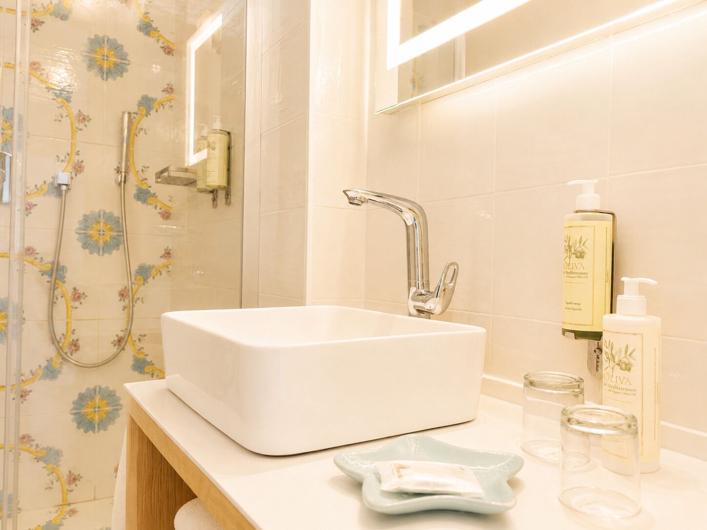 Un baño con un lavamanos blanco, una ducha con azulejos florales y artículos de aseo en la encimera.