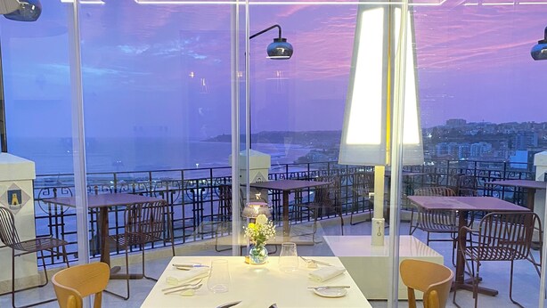 Un restaurant avec une table dressée pour deux personnes, des chaises et une vue sur le paysage urbain grâce à de grandes fenêtres.