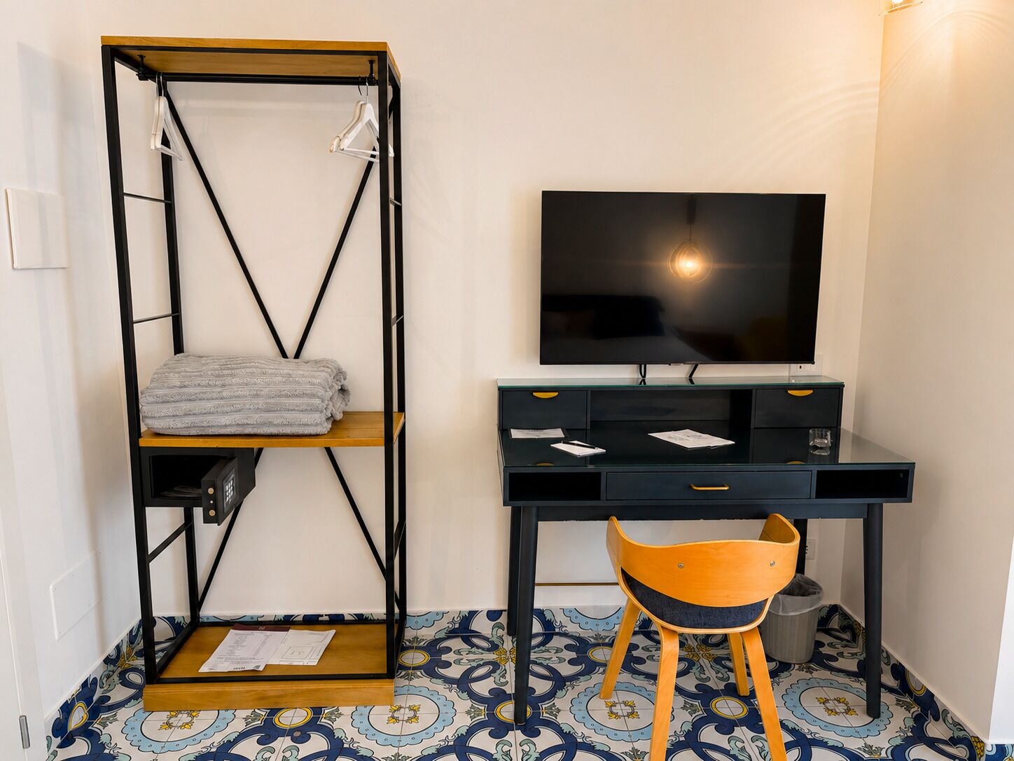 A room with a bed, a desk with a television, a chair, and a shelf with towels.