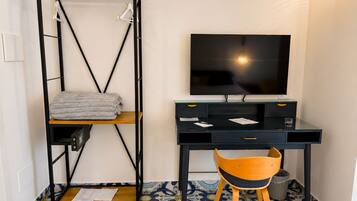 A room with a bed, a desk with a television, a chair, and a shelf with towels.