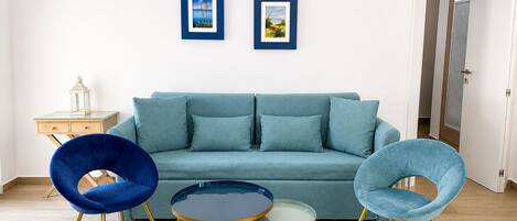 A living room with a teal sofa, two blue chairs, a small table, and two framed pictures on the wall.