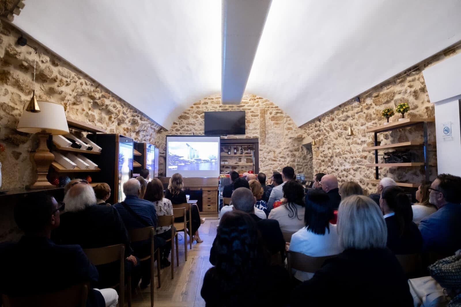 Una habitación con paredes de piedra, una pantalla de proyector mostrando un paisaje urbano, estantes de madera y un televisor.