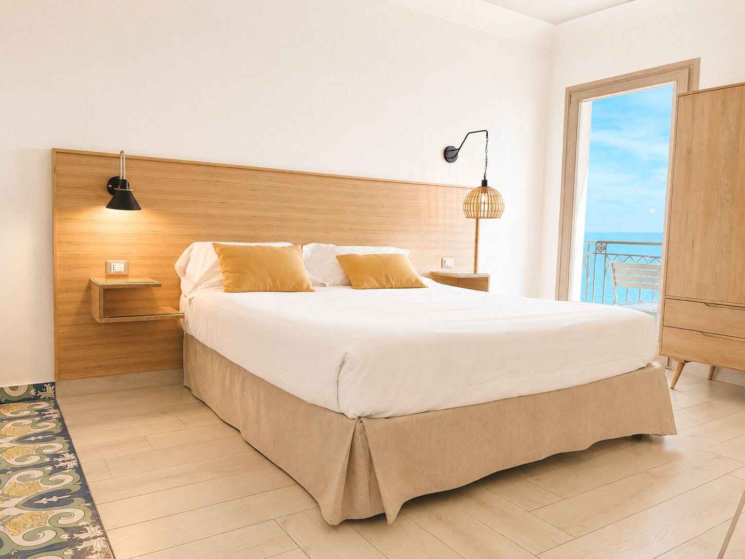 Una camera da letto con una testiera in legno, un letto con lenzuola bianche, due abat-jour, un comodino e vista mare tramite una porta finestra.