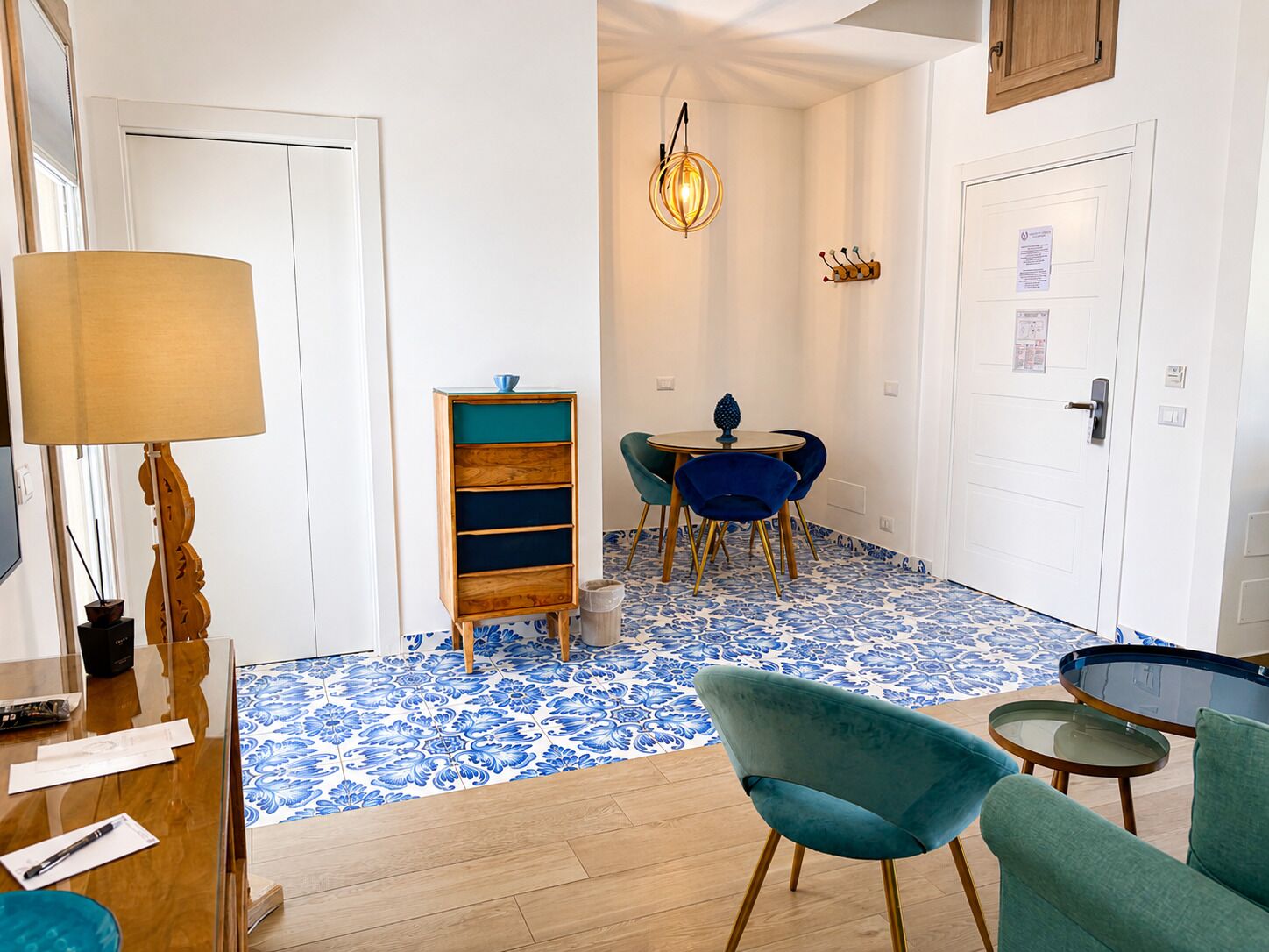 A room with a wooden desk, a lamp, a blue patterned floor, a dining table with chairs, and a small blue chair.