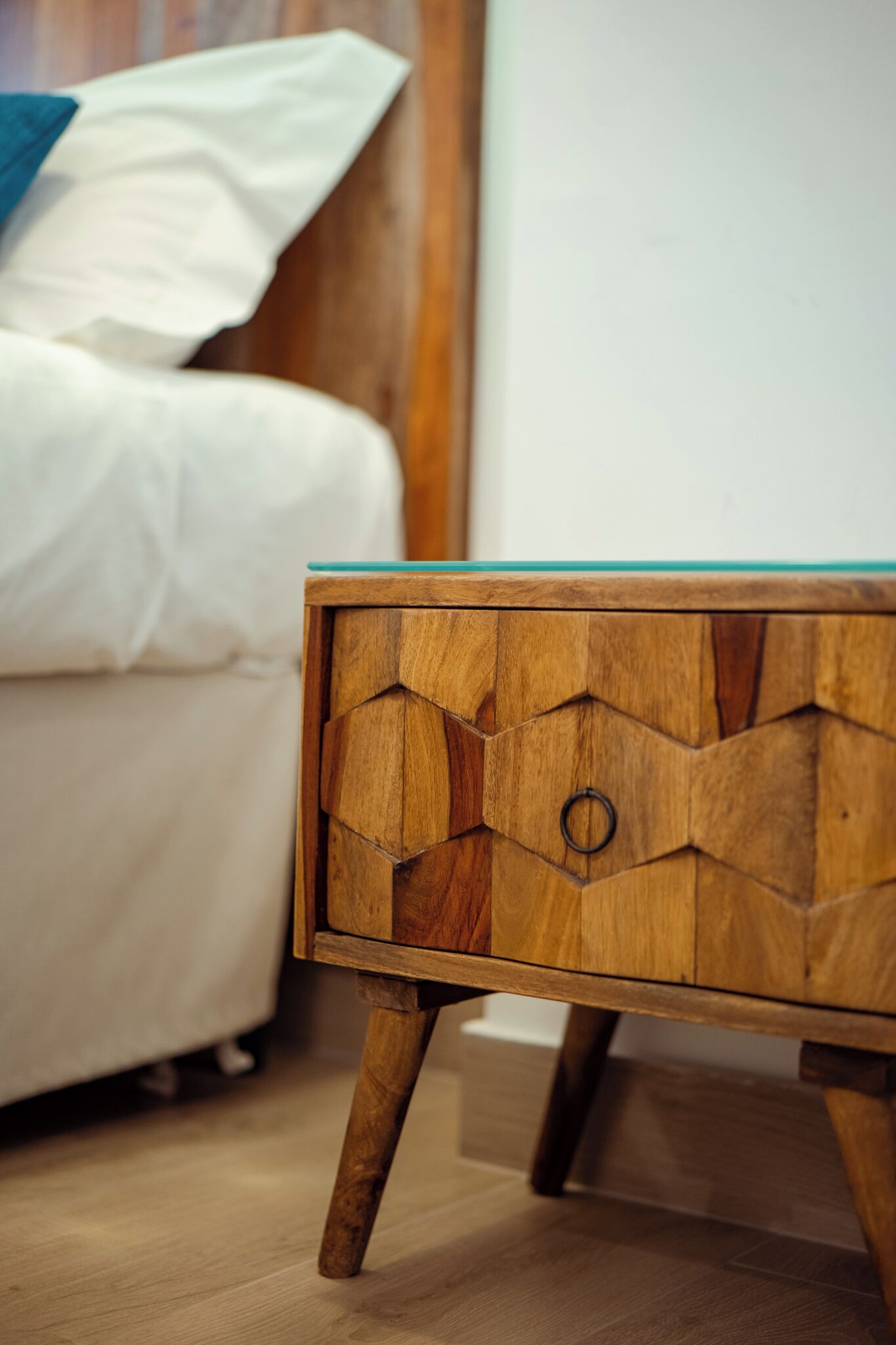 A wooden bedside table with a geometric design and a glass top, positioned next to a bed with white bedding and a wooden headboard.