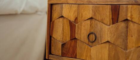 A wooden bedside table with a geometric design and a glass top, positioned next to a bed with white bedding and a wooden headboard.