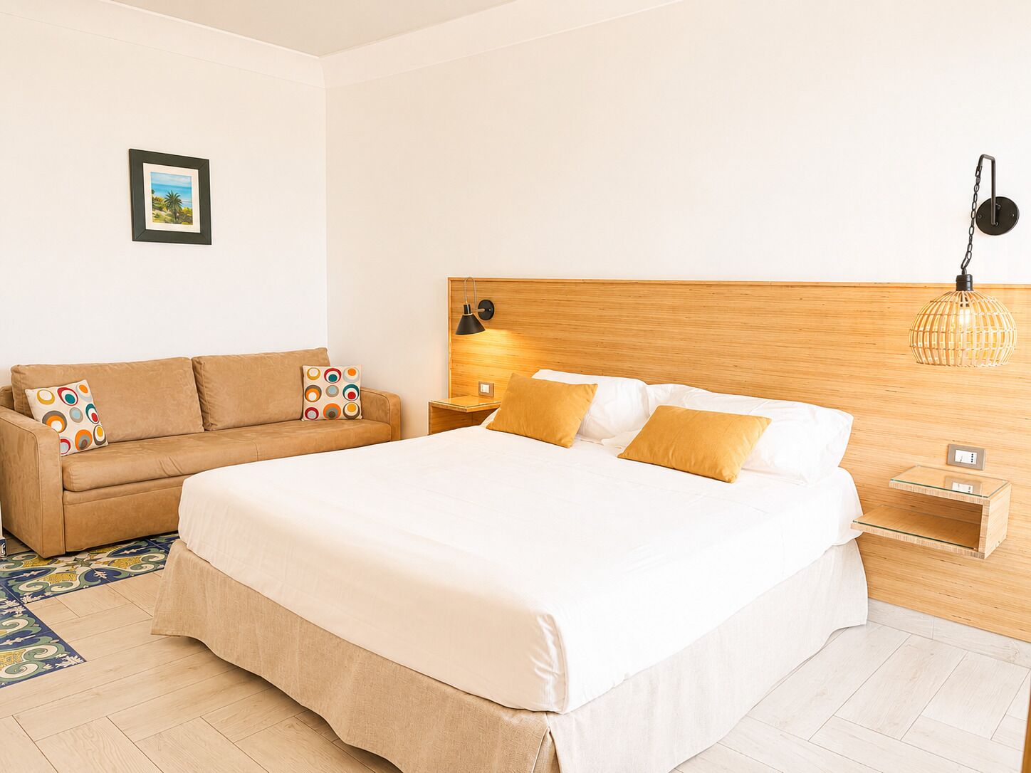 A hotel room with a bed, a sofa, a nightstand, a wall-mounted lamp, and a picture on the wall.