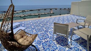 Un balcon avec un fauteuil suspendu, deux chaises et une table. Le balcon a un sol à motifs et offre une vue sur la mer.