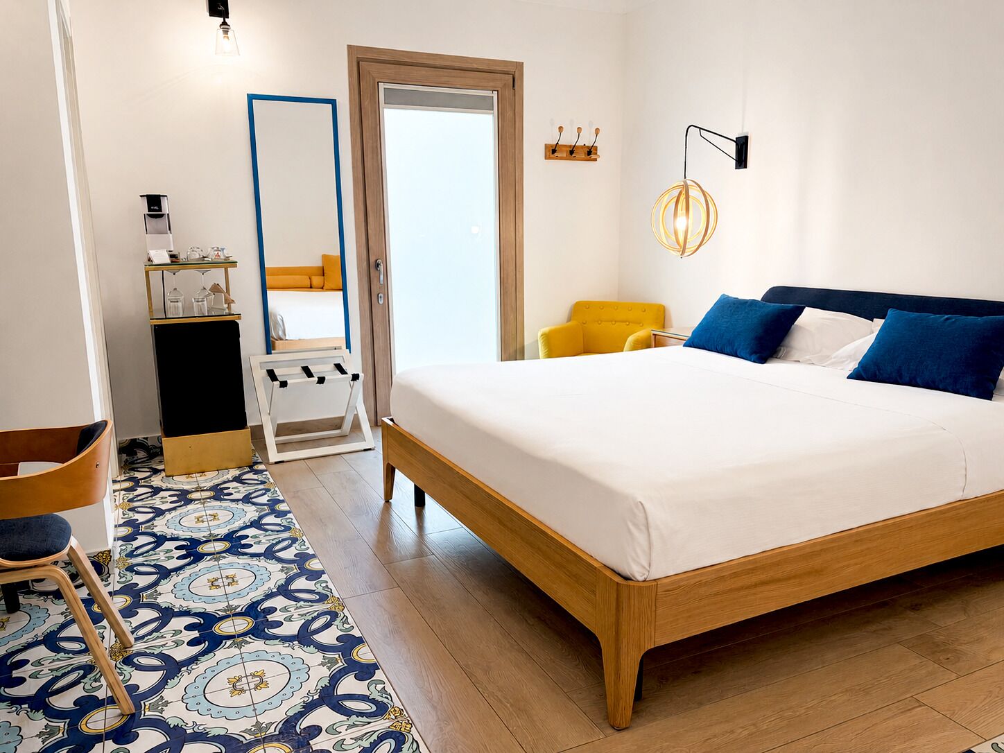 Habitación de hotel con una cama grande, una silla, una mesita, un espejo y una lámpara fijada en la pared.