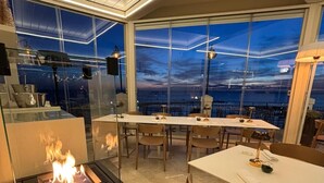 Un espace repas moderne avec une table centrale, des chaises et un barbecue intégré avec des flammes. De grandes fenêtres offrent une vue sur le paysage urbain au crépuscule.