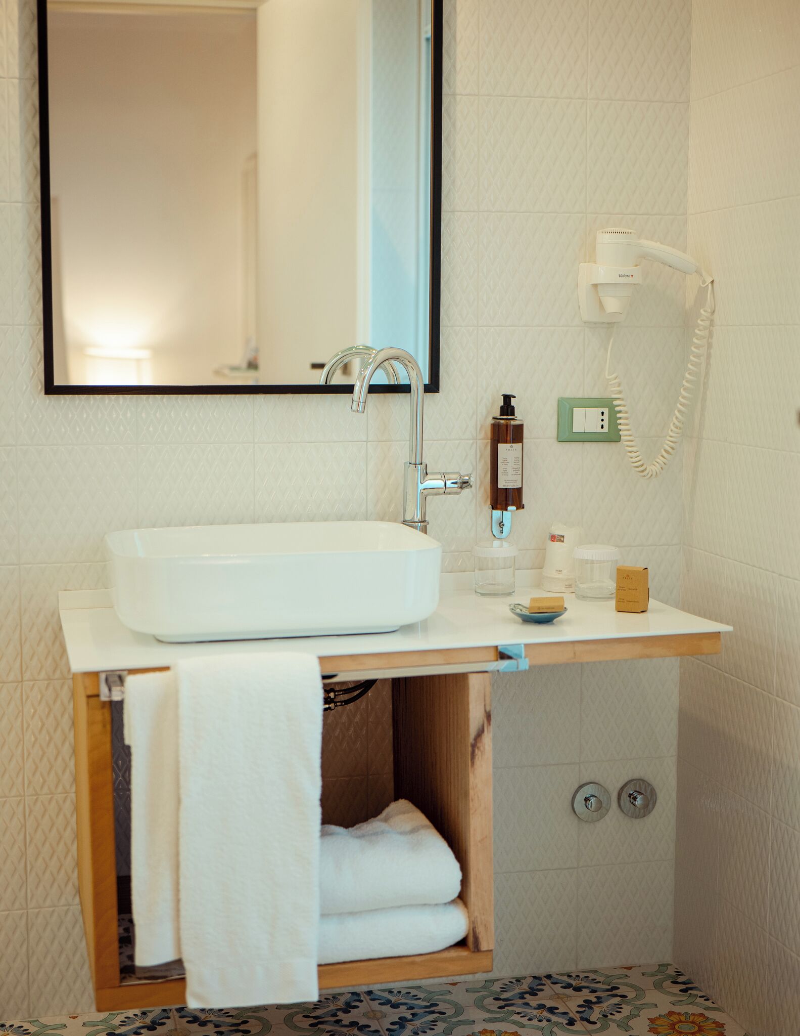 Une salle de bain avec un lavabo blanc, un miroir, un porte-serviettes, un sèche-cheveux et une petite étagère contenant des articles de toilette.