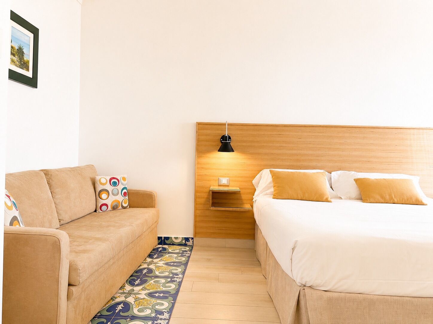 A hotel room with a bed, a sofa, a nightstand, and a wall-mounted lamp.