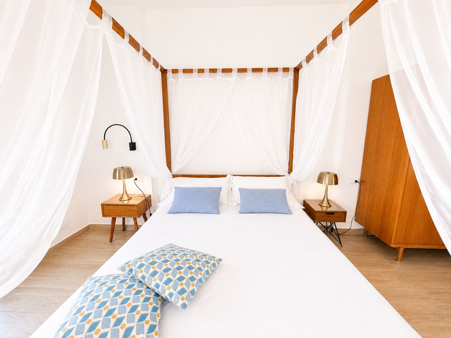 A four-poster bed with white drapes, two bedside tables with lamps, and a wooden wardrobe.