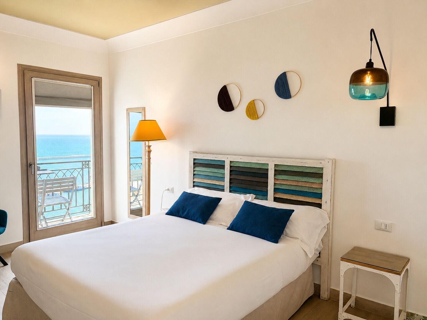 A hotel room with a bed, a nightstand, a lamp, and a view of the sea through a balcony door.