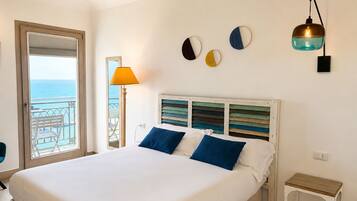A hotel room with a bed, a nightstand, a lamp, and a view of the sea through a balcony door.
