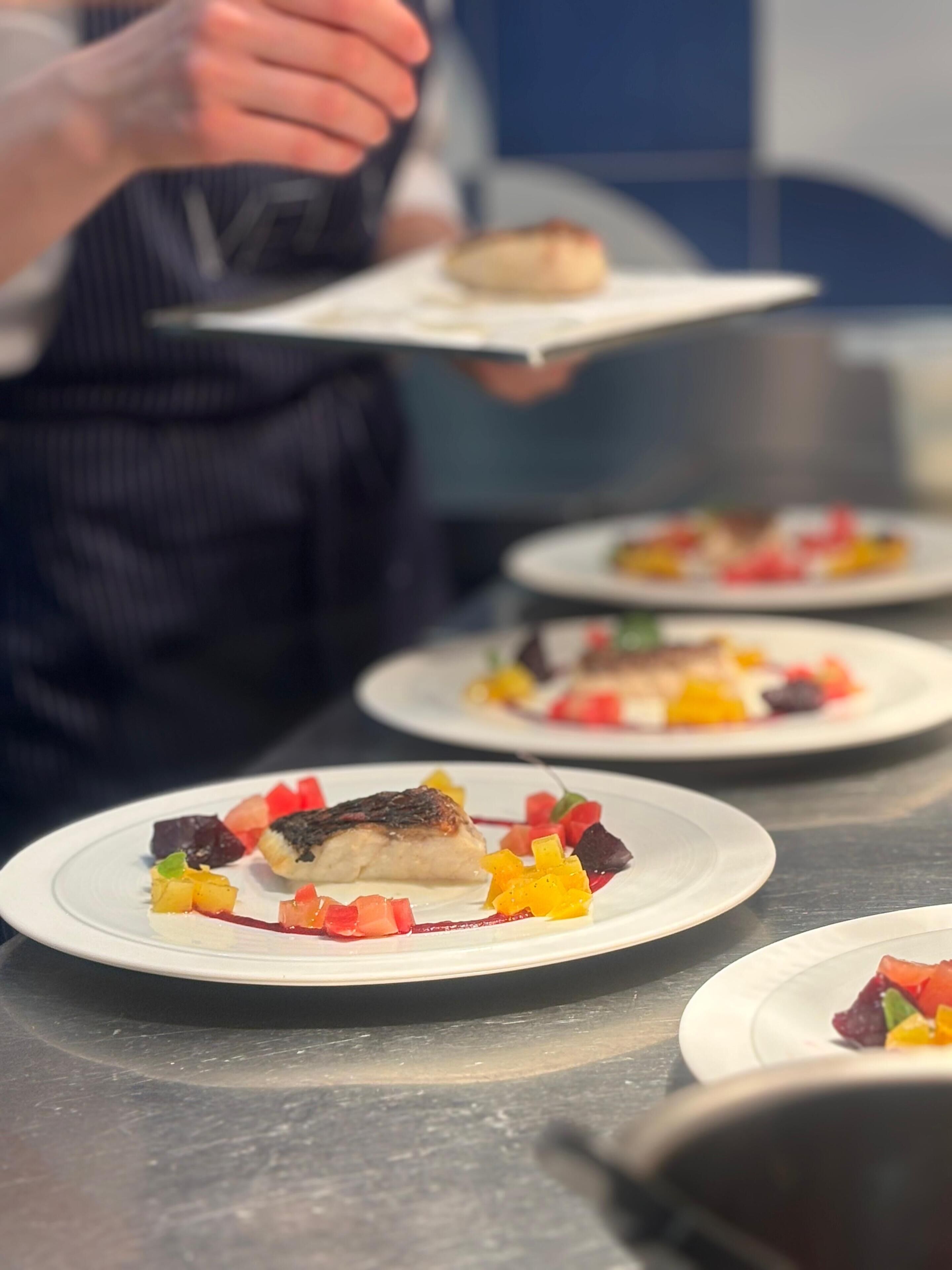Un chef tenant une assiette contenant un plat de poisson, entouré d’autres assiettes sur lesquelles se trouvent divers aliments.