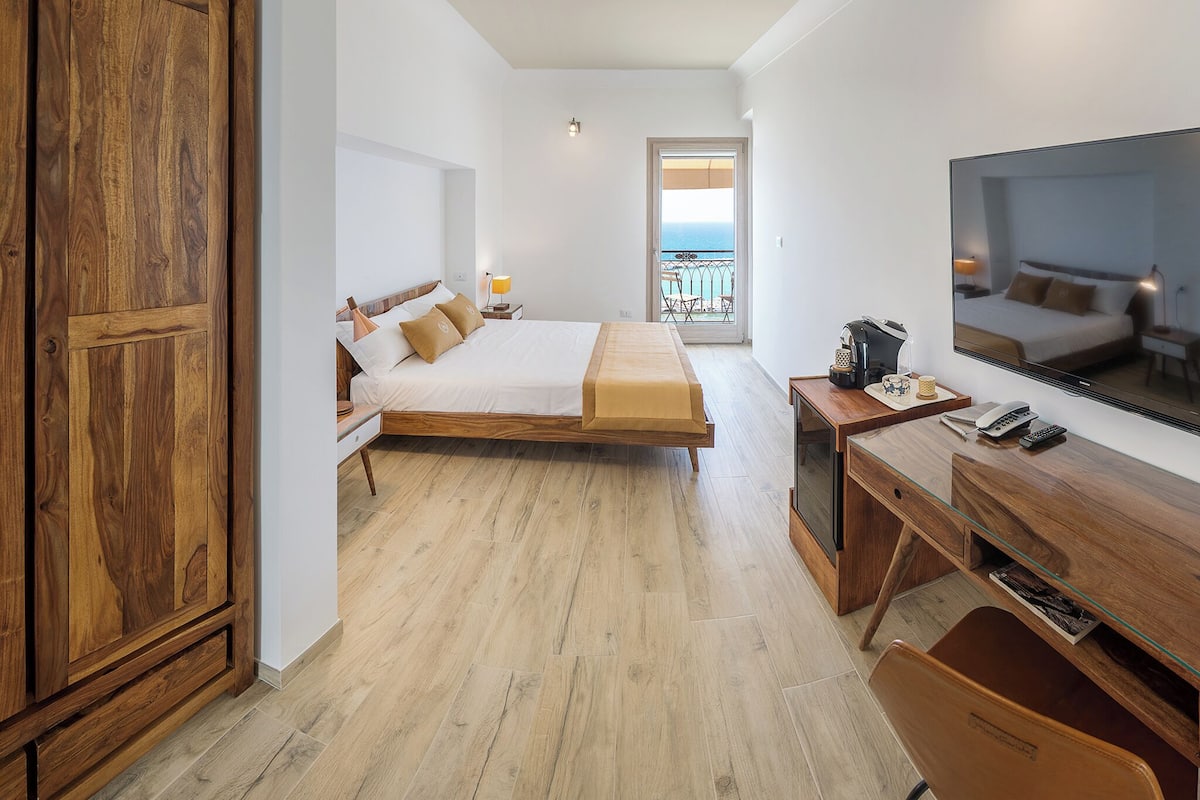 Ein Schlafzimmer mit Bett, Holztisch, Stuhl, Fernseher und einem Balkon mit Blick.