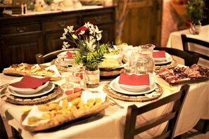A dining table set with various dishes, including bread, pastries, and a meat platter, with a vase of flowers and a pitcher of water. - Country House S'Ozzastru (Dorgali)