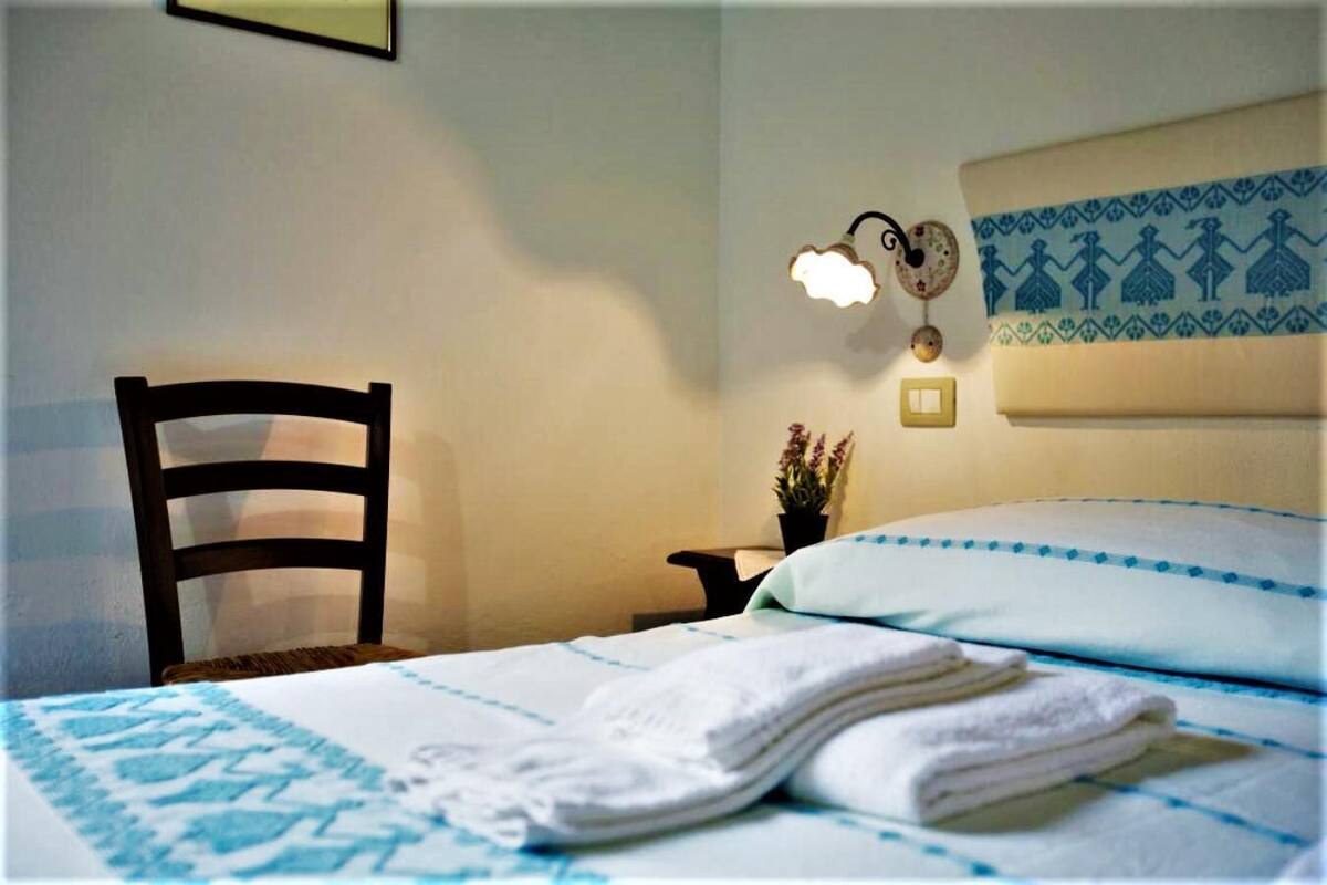 A hotel room with a bed, a chair, a nightstand, and a wall-mounted light fixture.