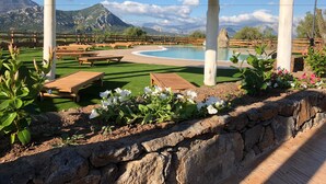A pool area with wooden benches and a stone wall, surrounded by mountains and a clear sky. - Country House S'Ozzastru (Dorgali)