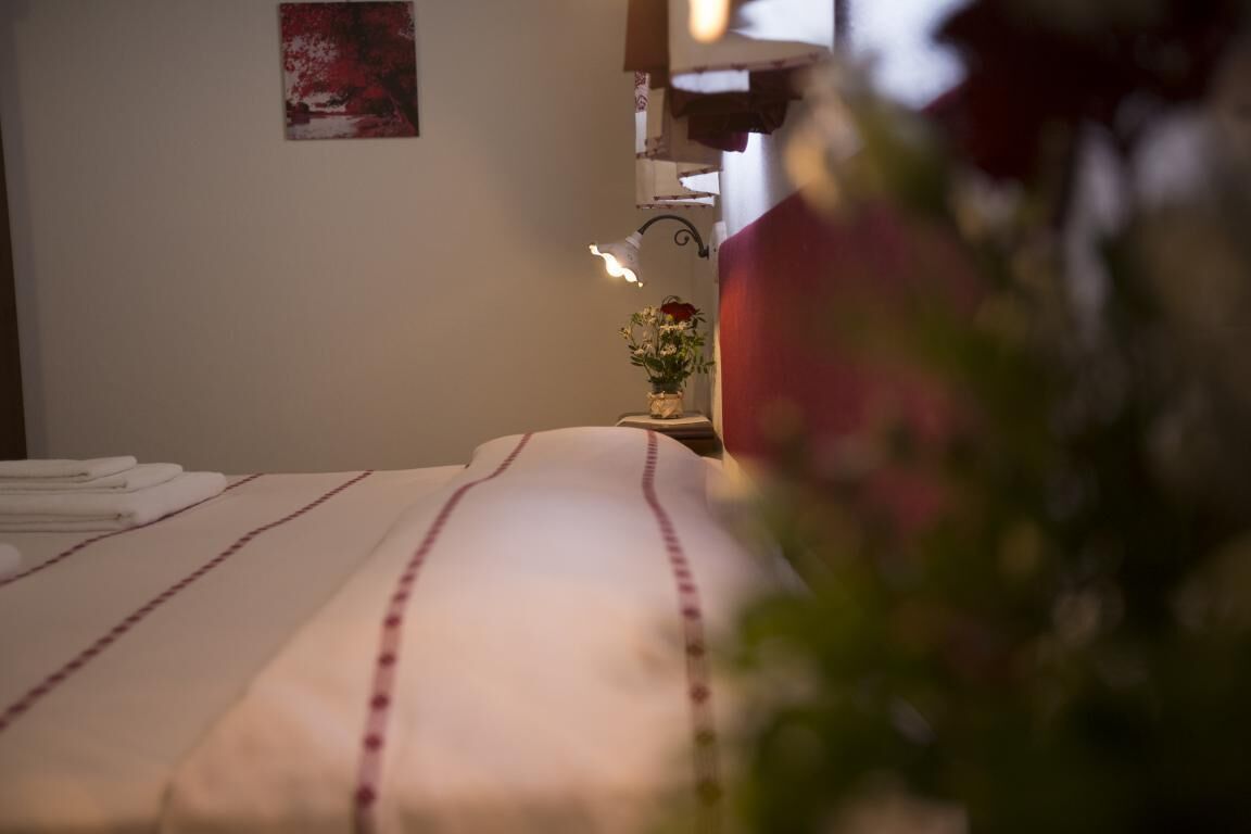 Ein ordentlich bezogenes Bett mit einer weiß-rot gestreiften Tagesdecke, ein kleiner Nachttisch mit Blumenvase und eine an der Wand befestigte Lampe.