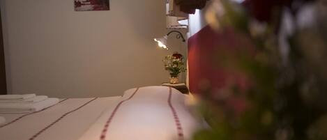 Ein ordentlich bezogenes Bett mit einer weiß-rot gestreiften Tagesdecke, ein kleiner Nachttisch mit Blumenvase und eine an der Wand befestigte Lampe.