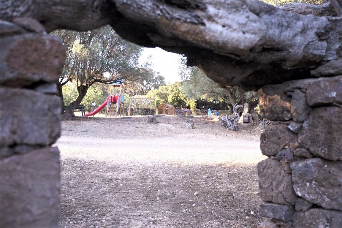 A park with a playground, including a slide and climbing structures, surrounded by trees and stone walls.