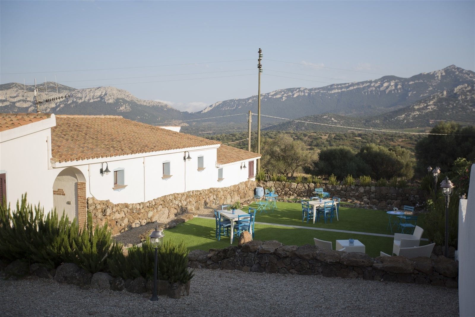 Un edificio blanco con tejado de tejas, paredes de piedra y base de piedra. Cuenta con un patio con mobiliario exterior y vistas a montañas al fondo.