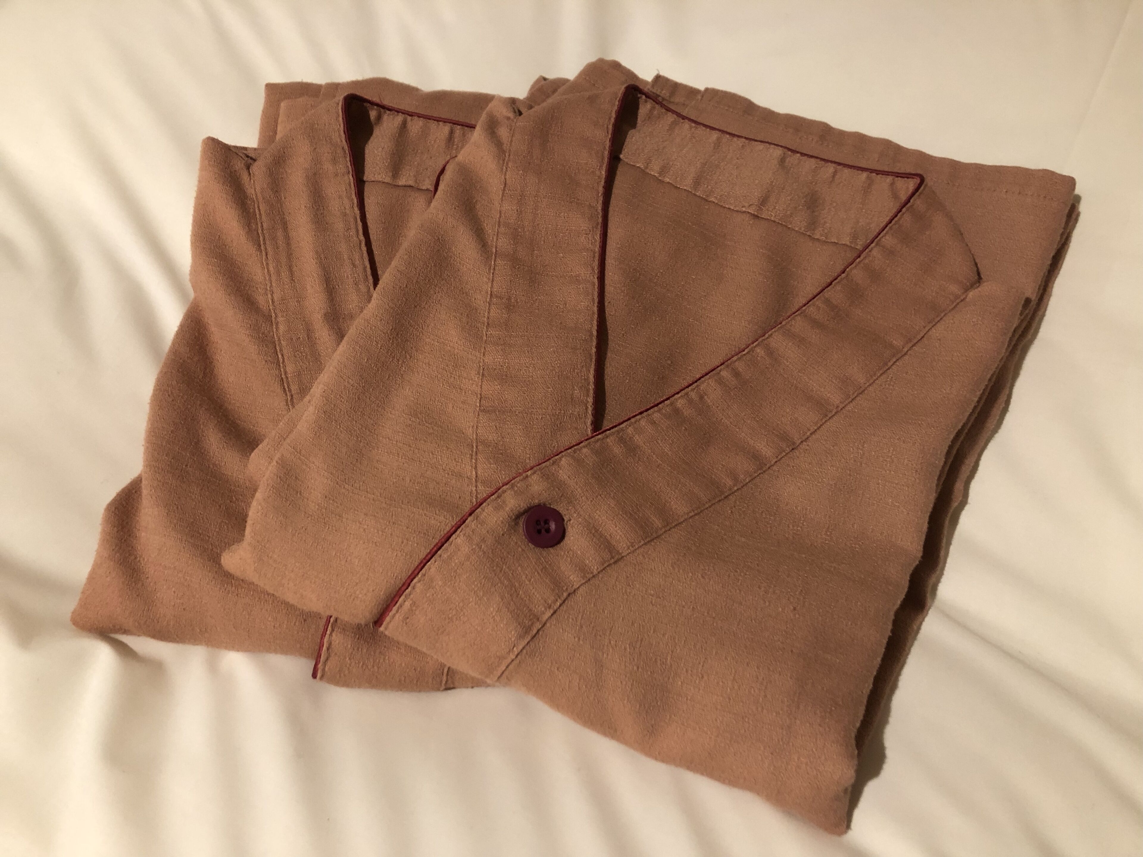 Una camisa marrón bien doblada con un botón y un detalle rojo sobre una cama blanca.