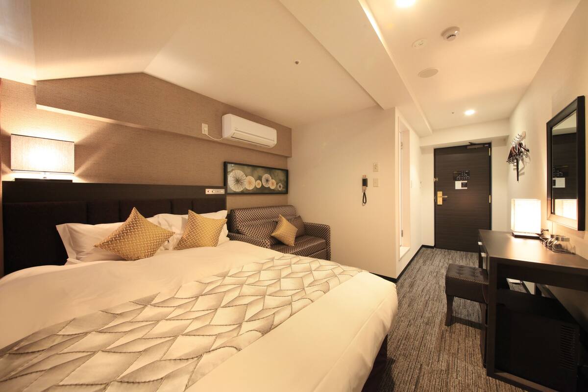 Una habitación de hotel con una cama grande, un escritorio, una silla, un equipo de aire acondicionado y una puerta que da a otra habitación.