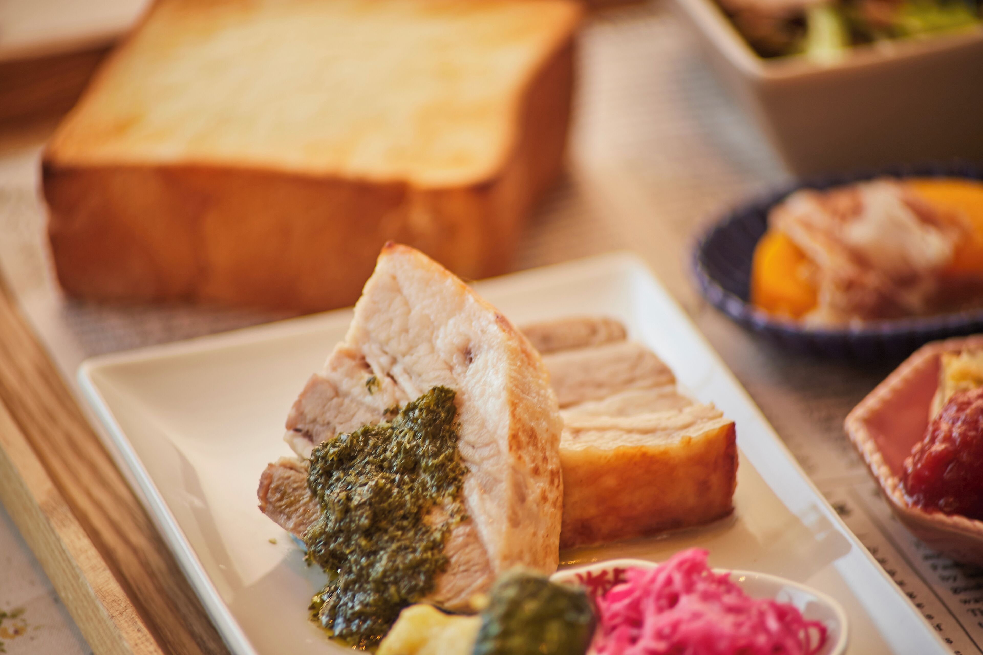 Une assiette avec des tranches de viande et une sauce verte, une miche de pain et un petit bol de fruits sur une table.