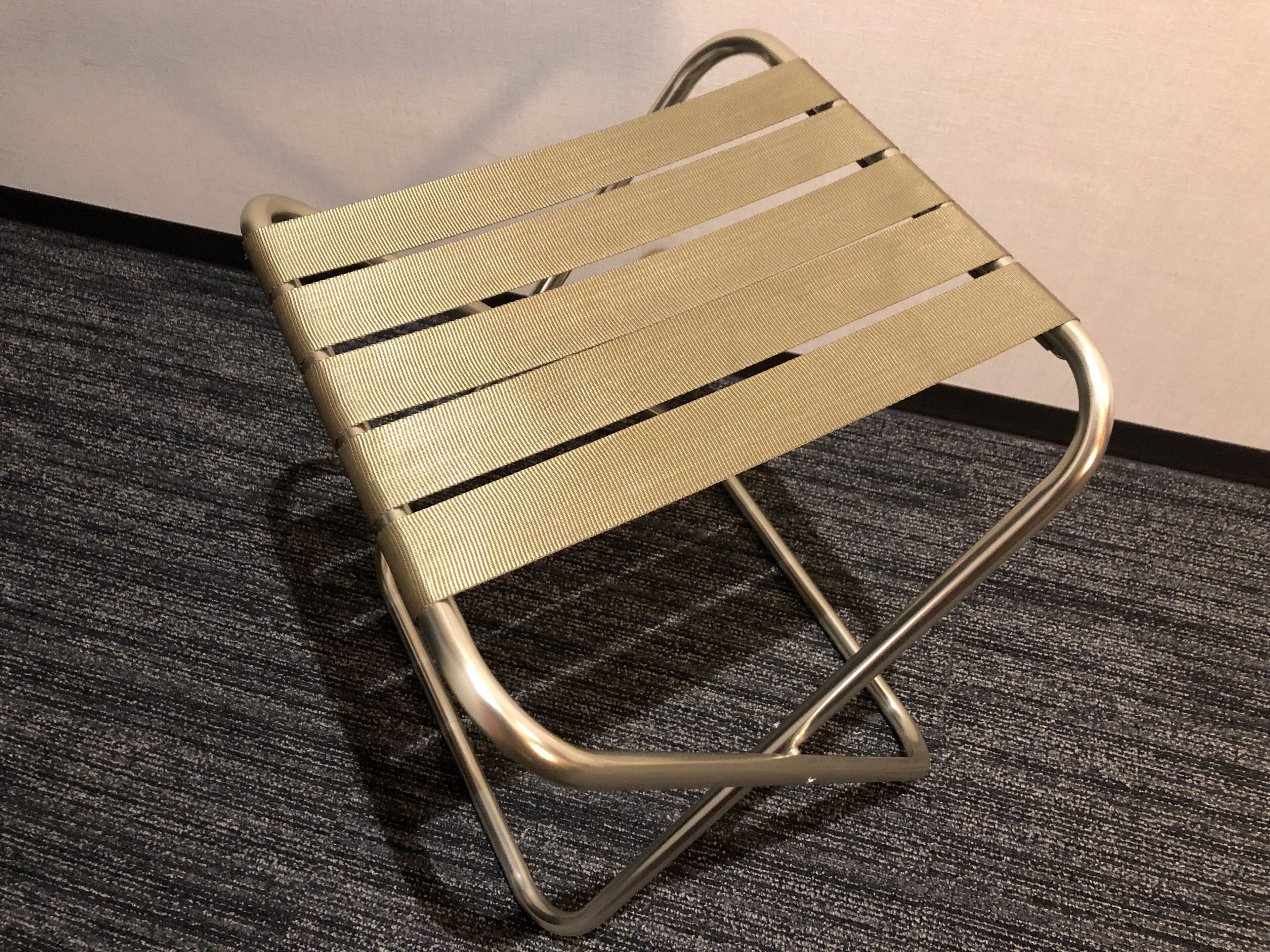 Een metalen kruk met een geweven zitting en rugleuning, op een getextureerd vloerkleed geplaatst.