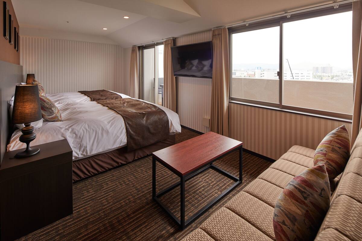 Habitación de hotel con cama, sofá, mesa de centro, televisor y ventana con vista a la ciudad.