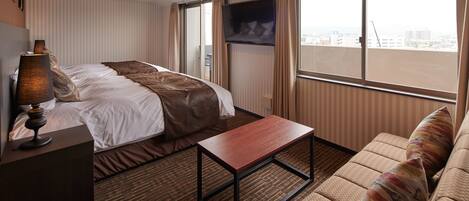 A hotel room with a bed, a sofa, a coffee table, a TV, and a window with a city view.