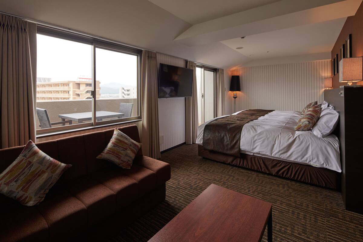 Habitación de hotel con una cama grande, un sofá, un televisor y un balcón con vistas a edificios.