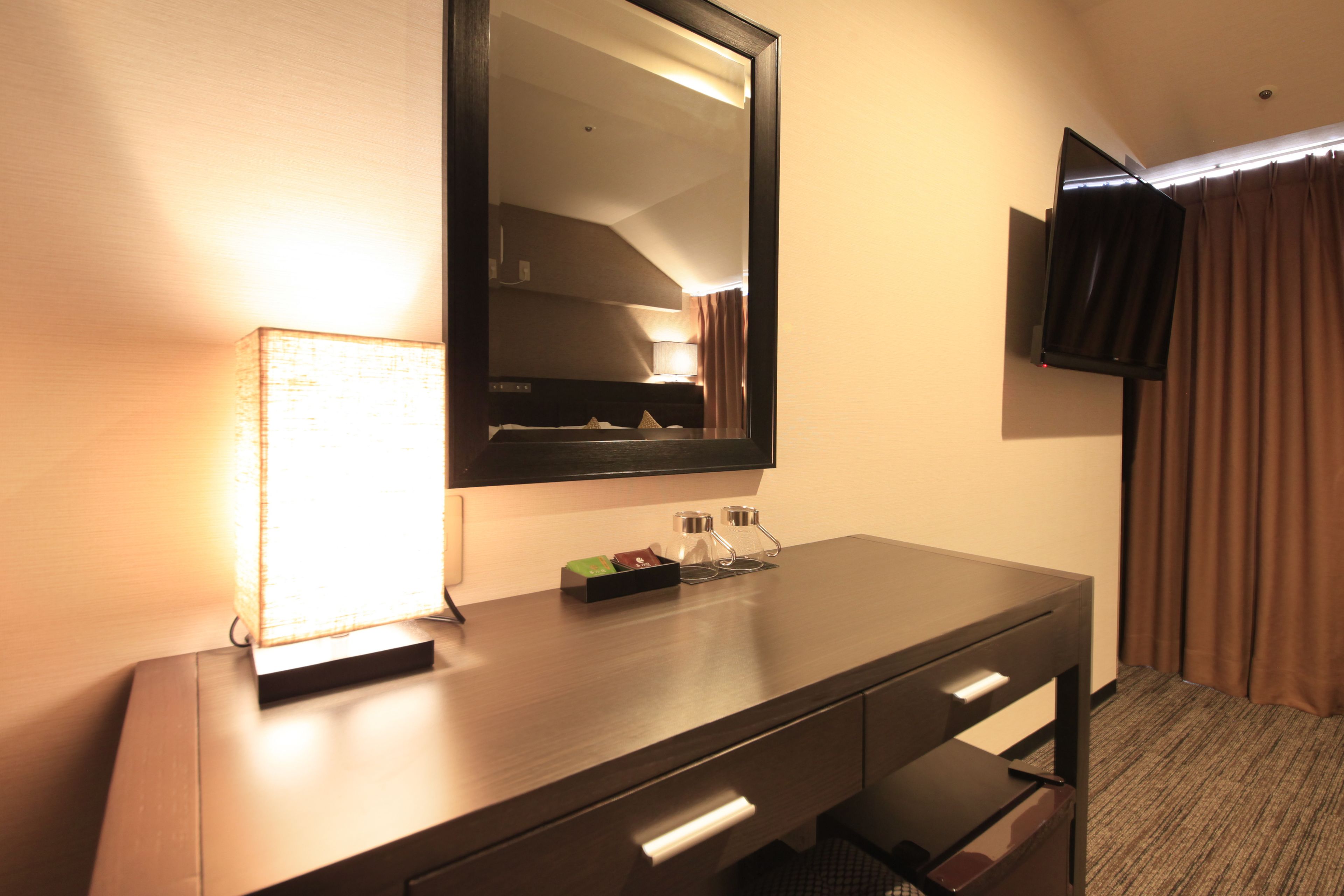 A hotel room with a wooden dresser, a mirror, a flat-screen TV mounted on the wall, and a bed with brown curtains.