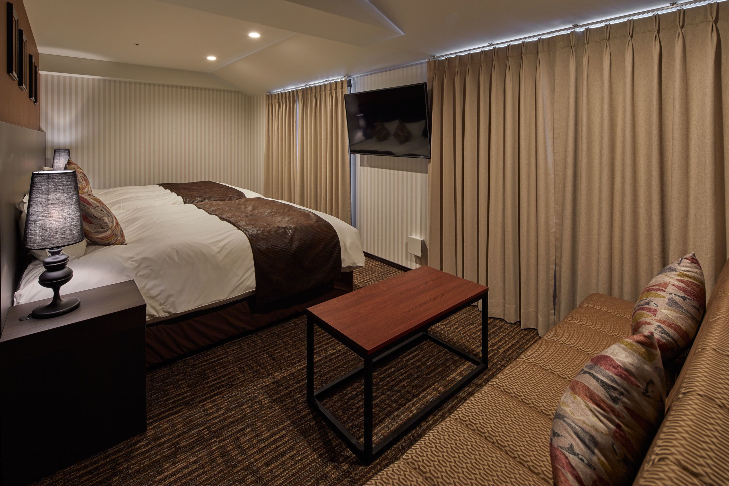 A hotel room with a bed, a bedside table with a lamp, a wooden bench, a sofa, and a TV mounted on the wall.