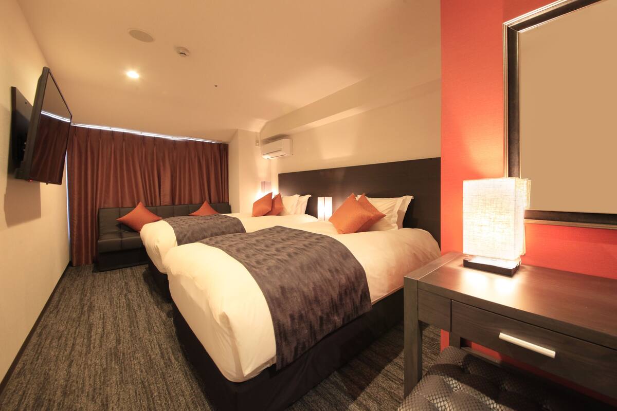Habitación de hotel con dos camas, un escritorio, una silla, un televisor montado en la pared y un equipo de aire acondicionado.