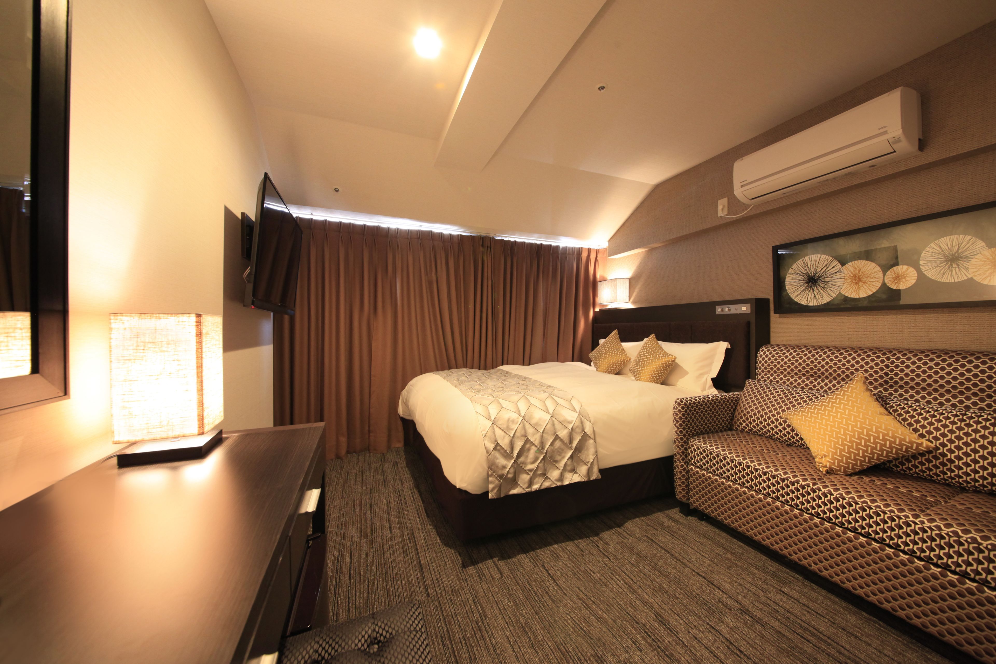 A hotel room with a bed, a sofa, a desk, a TV, and an air conditioning unit.