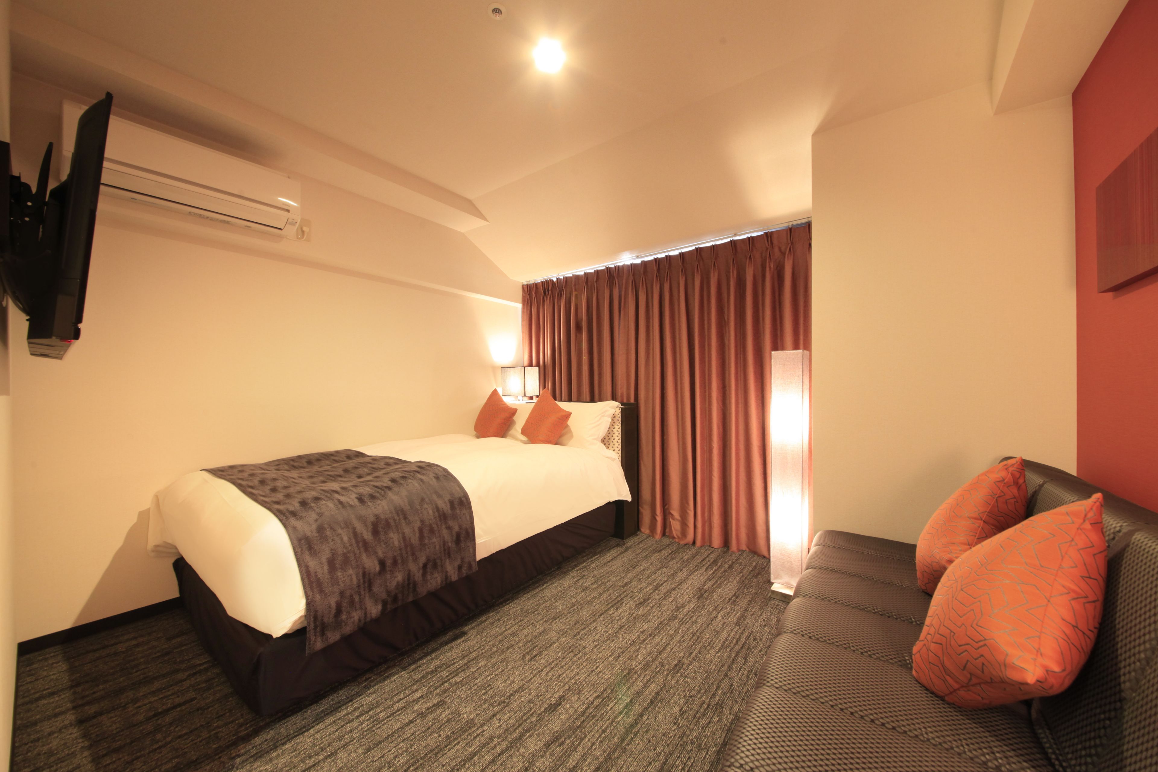 Une chambre d’hôtel avec un lit, un canapé, un téléviseur fixé au mur et une fenêtre avec des rideaux.
