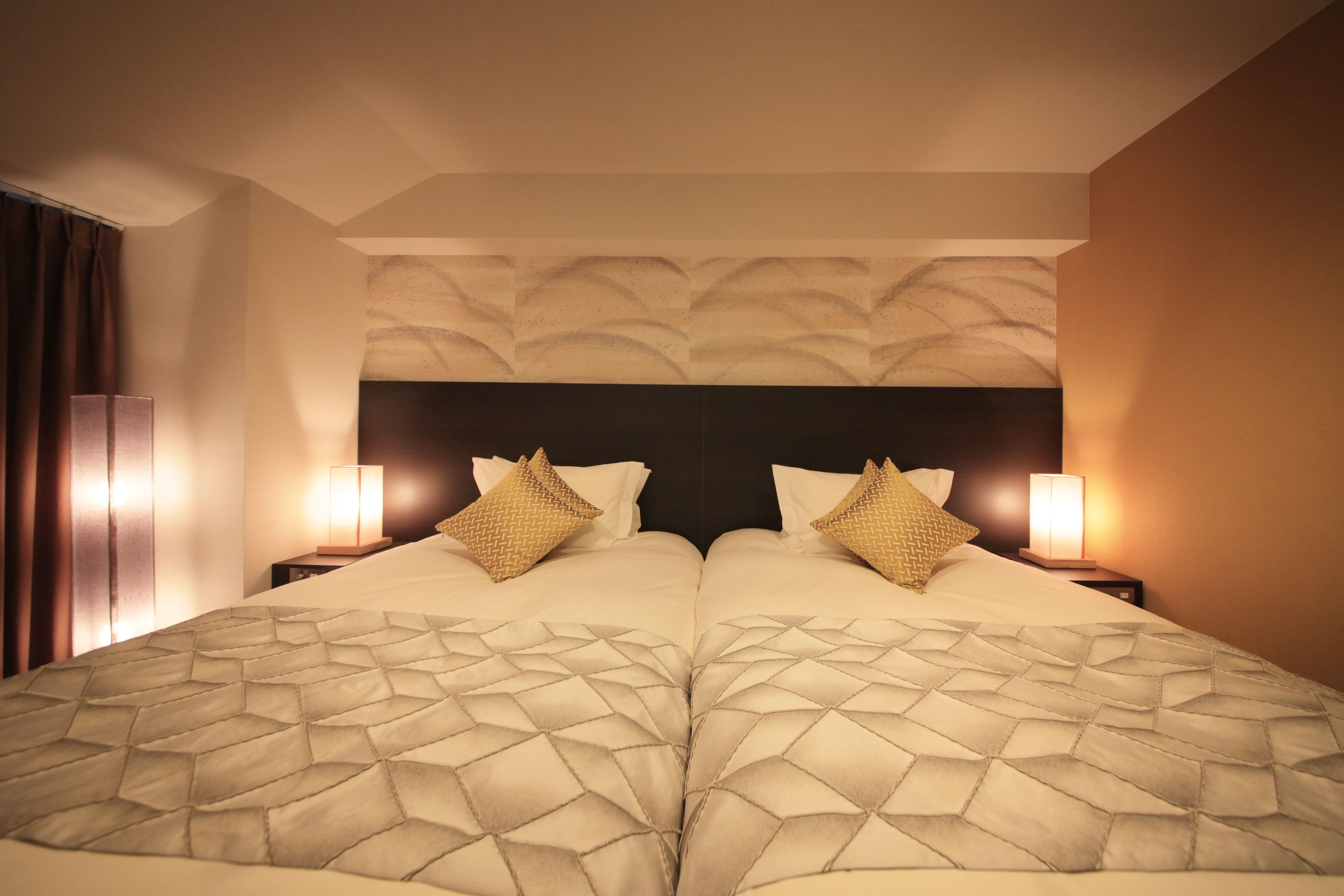 A double bed with a geometric patterned bedspread, two bedside lamps, and a textured wall headboard.