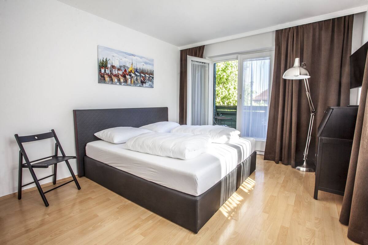 A modern hotel room with a bed, a desk, a chair, and a painting on the wall.