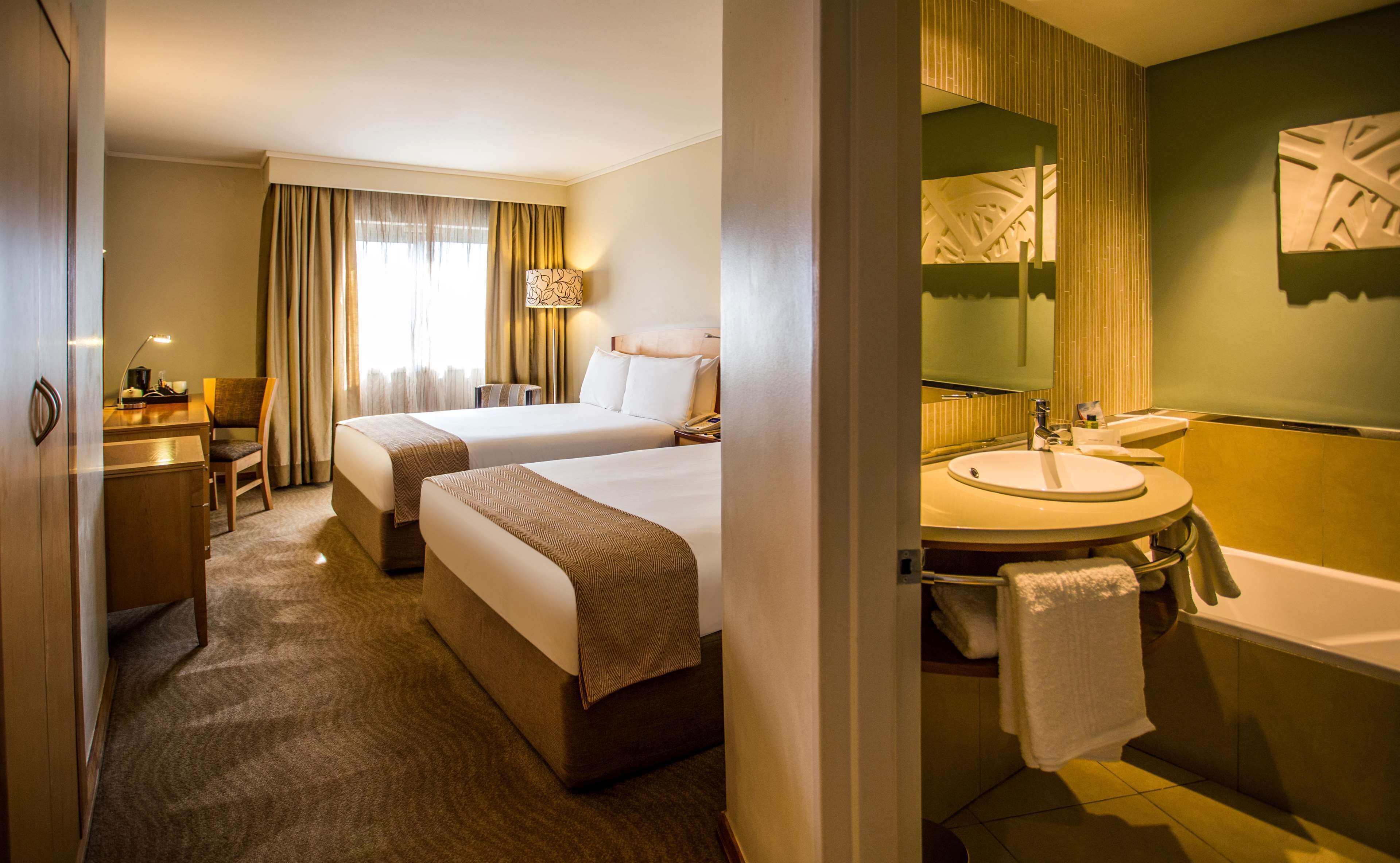 Une chambre d’hôtel avec deux lits, un bureau, une chaise, une lampe, et une salle de bain équipée d’un lavabo et d’une baignoire.