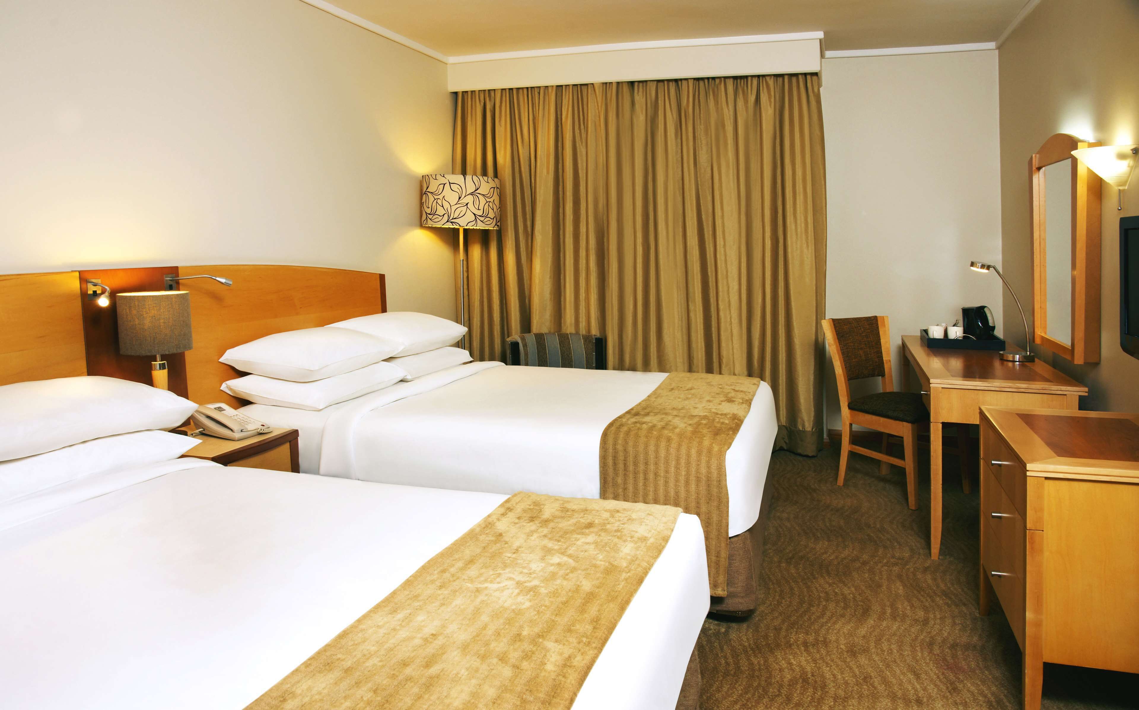 A hotel room with two beds, a desk, a chair, a lamp, and a large window with curtains.