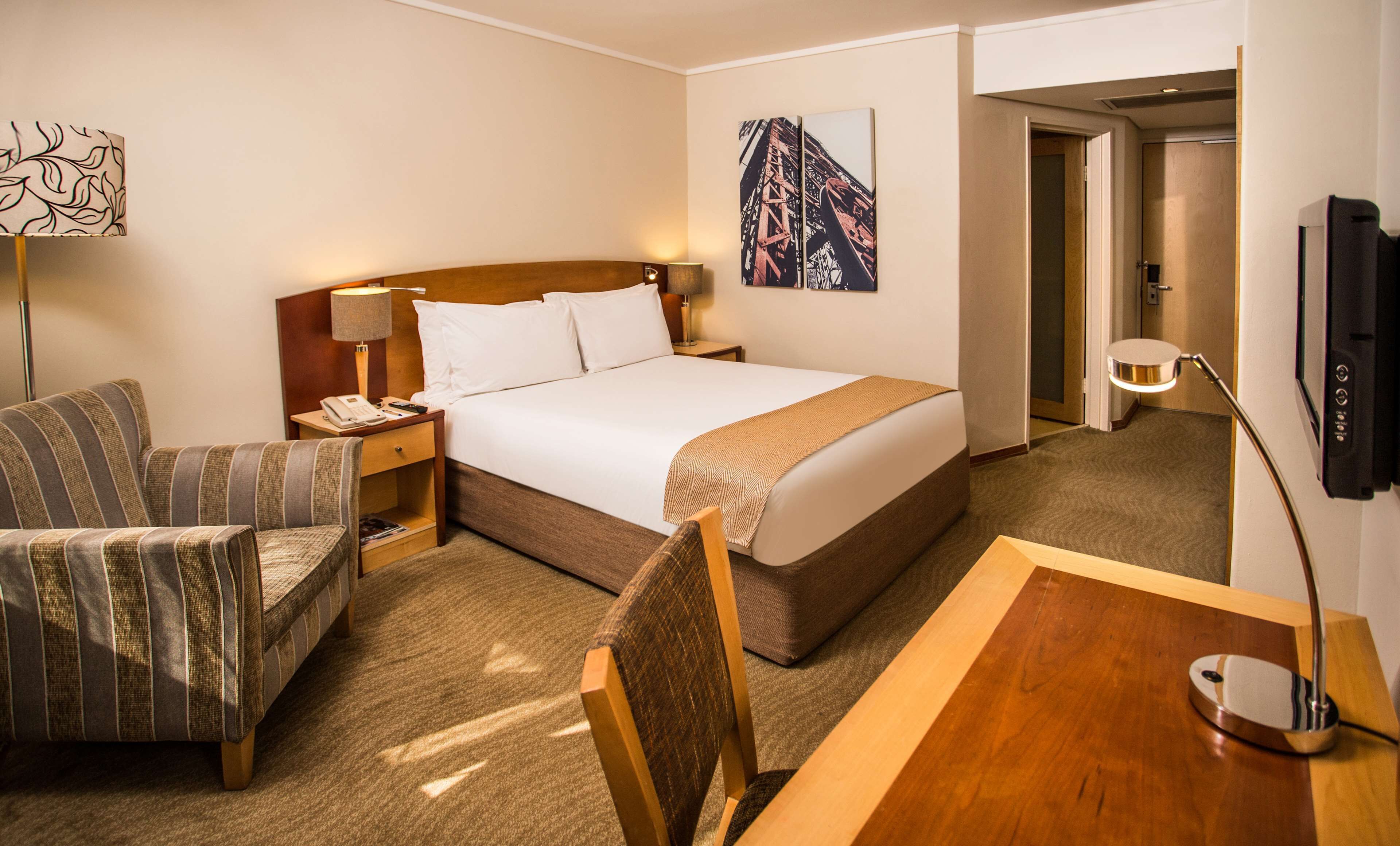 Ein Hotelzimmer mit einem Bett, einem Schreibtisch, zwei Sesseln, einem Nachttisch, einer Lampe, einem Fernseher und Bildern an den Wänden.