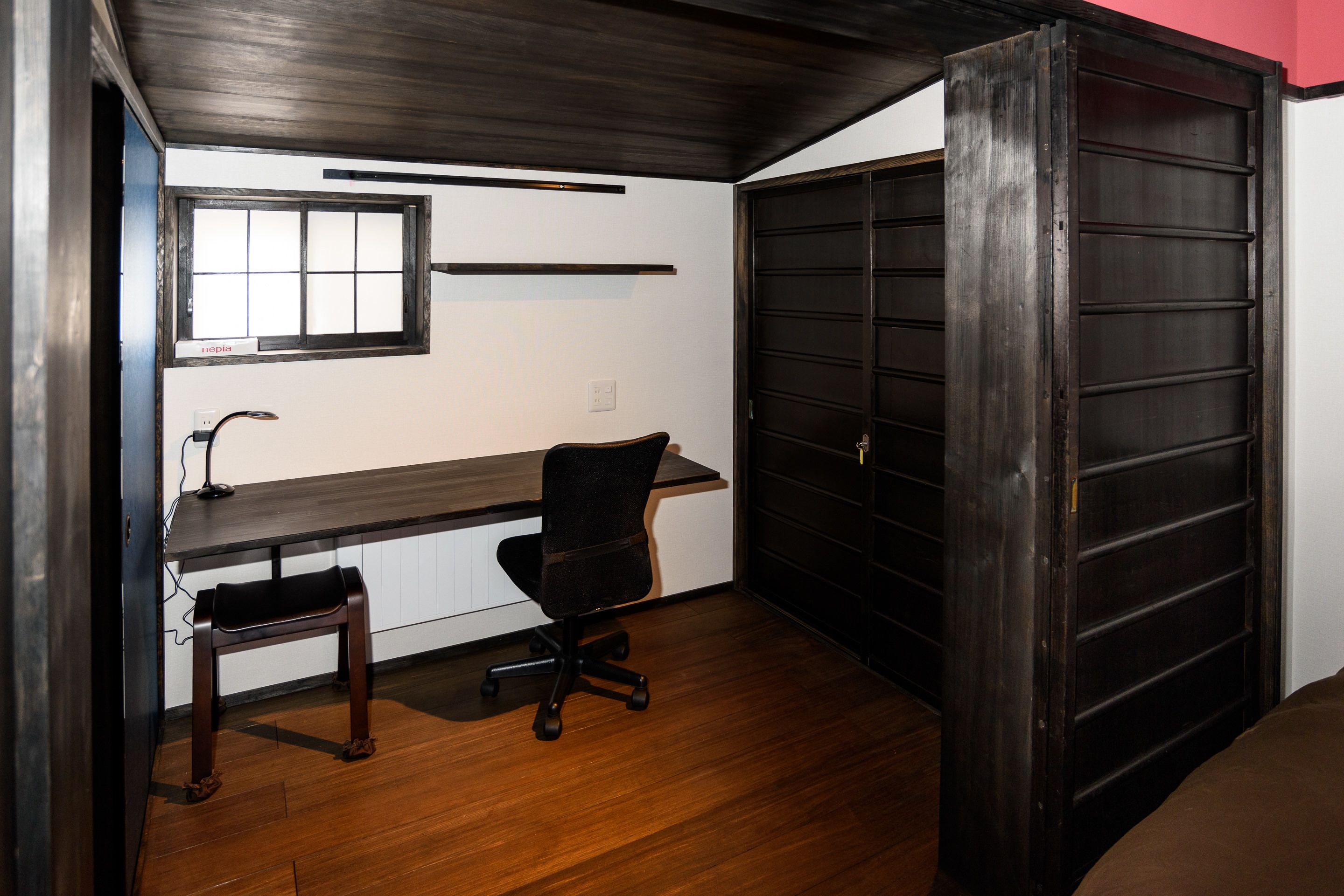 Een kamer met een houten bureau, een zwarte bureaustoel, een raam en een donkerhouten deur.