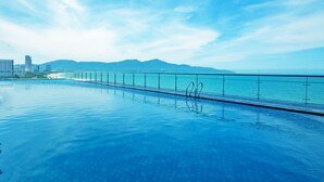 Una piscina con barandilla de vidrio, vista a la playa y montañas al fondo.