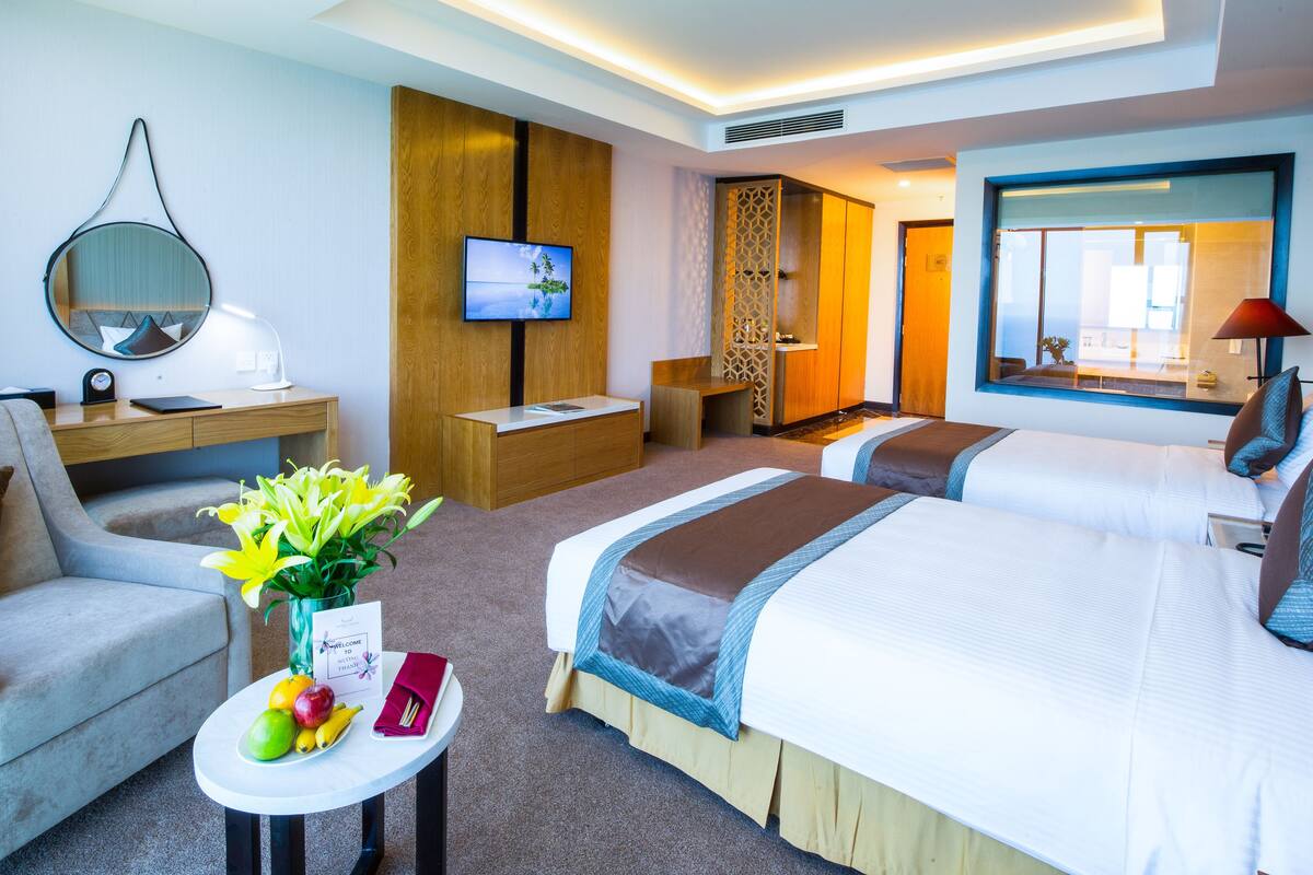 A hotel room with two beds, a desk, a chair, a TV, and a small table with fruit and a welcome sign.