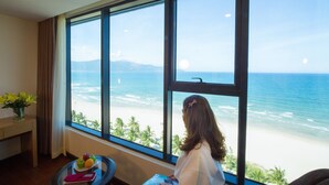 Una habitación con un ventanal grande que da a la playa. Hay una mesa con una bandeja de frutas y un florero con flores.