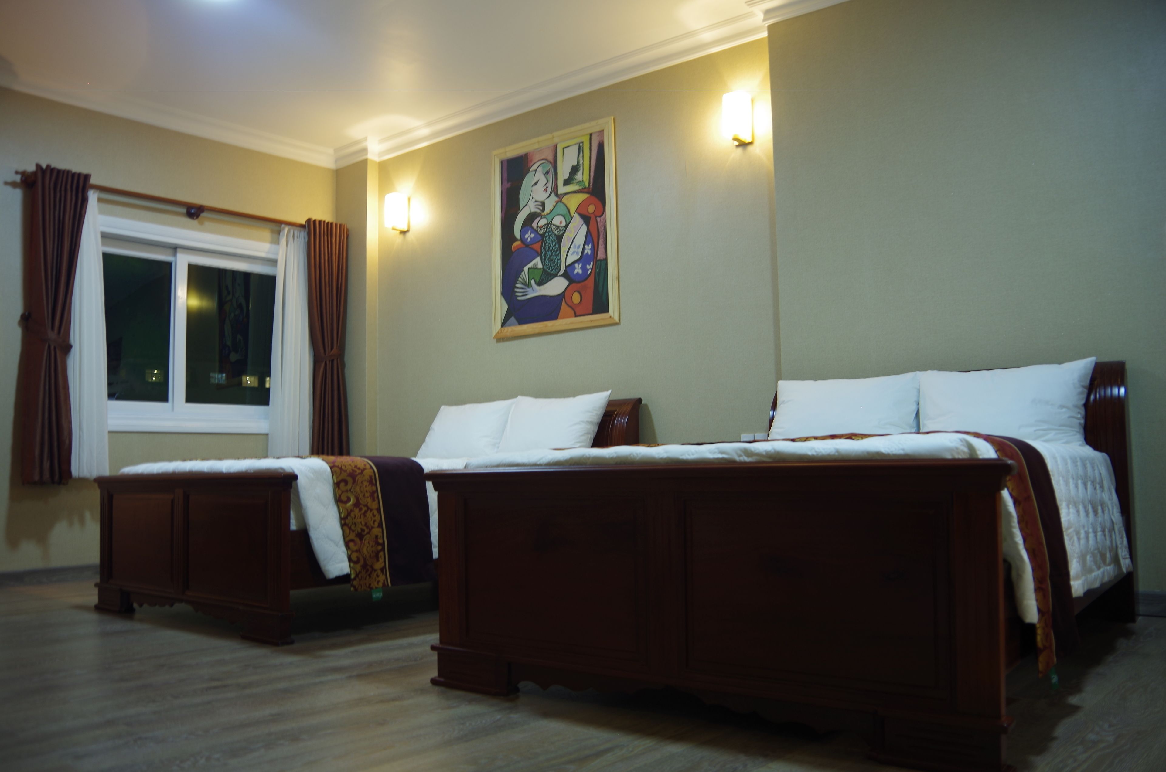 A hotel room with two wooden beds, a window with curtains, a painting on the wall, and a light fixture.